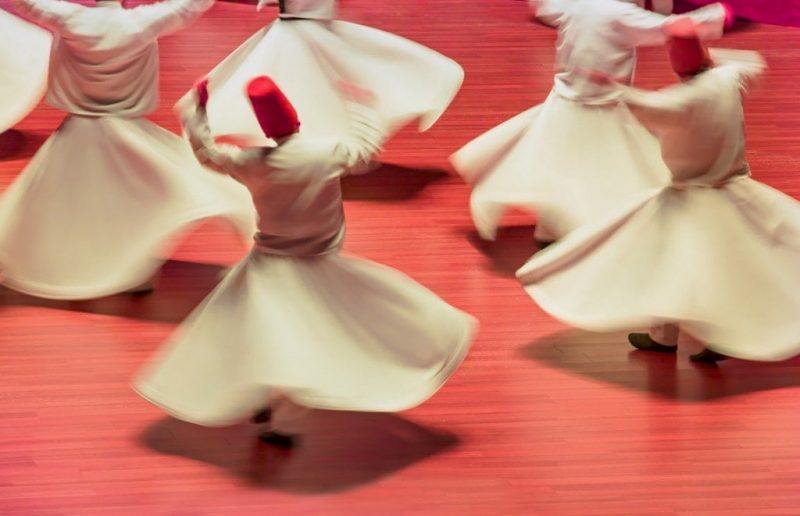 looking down at a red floor with five men in long white robes with flared white skirts and tall red fez style hats spinning around very fast so that the skirts flare out. 