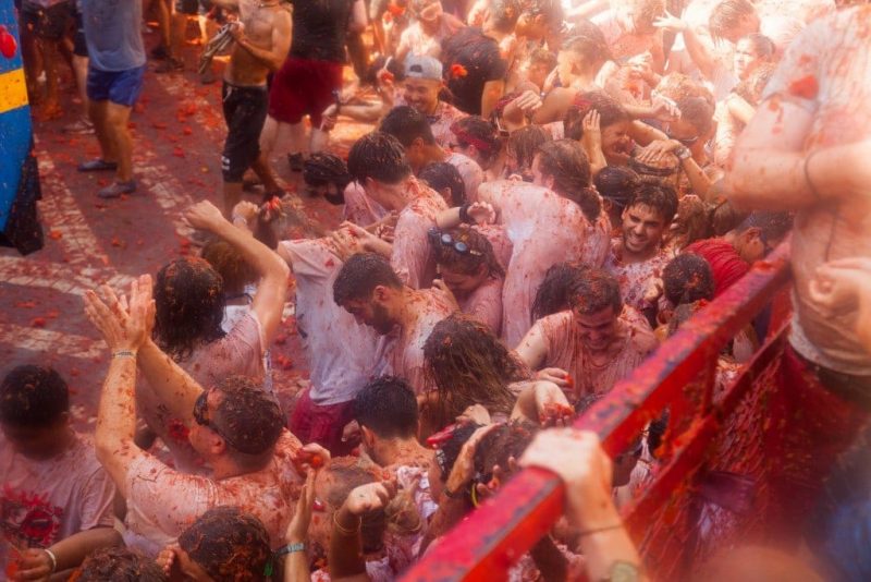 crowd of people at la Tomatina festival in Spain covered in red tomato pulp and sitting on the floor next to a red metal barrier