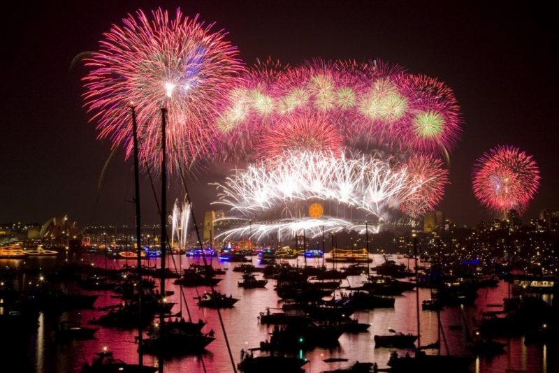 Fireworks in the night sky above Sydney Harbour which is filled with hundreds of boats in silhouette against the red glow of the fireworks reflected on the water