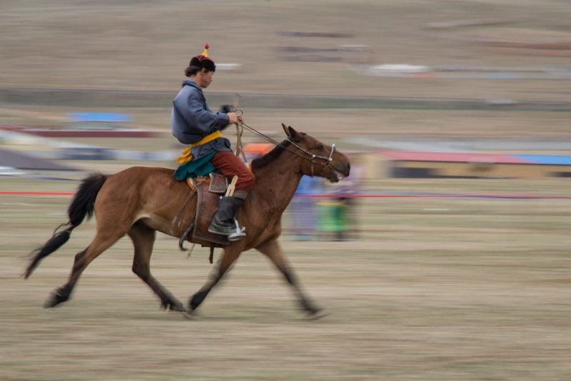Mongolian man wearing traditional dress with a blue shirt and brown trouses and a small red and yellow hat riding a brown horse very fast with the background and ground blurred due to the motion