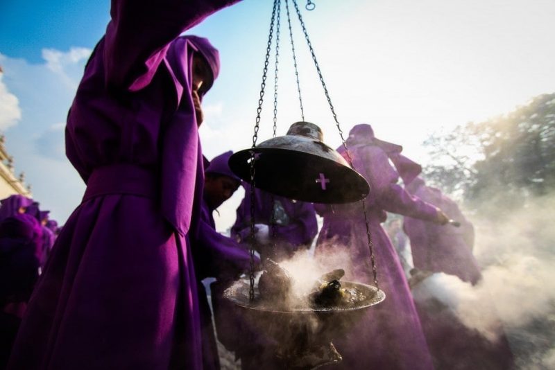 taken from a low angle looking up at a group of people wearing dark purple hooded robes at the Semana Santa procession in Guatemala, the person closest to the camera is holding a large metal censer in metal chains filled with burning cole and wafting smoke around the group