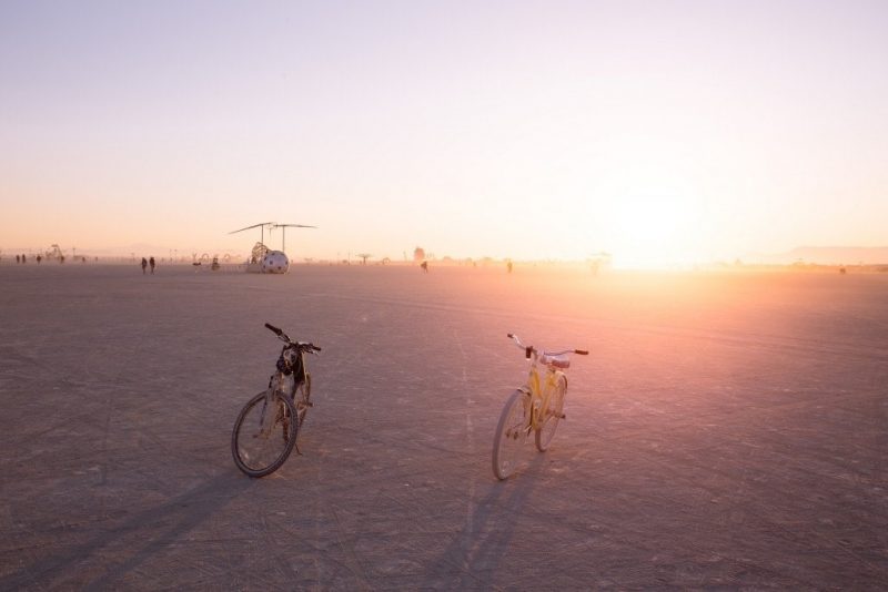 two bikes, one grey one yellow, parked on the white sand of the desert at Burning Man festival in Nevada with a washed-out sunset sky behind