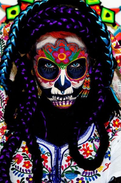close up of a woman's head and shoulders wearing a Dia de Muertos costume at the festival in Oaxaca. Her face is painted like a skull with a brightly coloured pattern around the eyes and forehead and a pink flower in the middle of her forehead, and she is wearing thick black and purple plaits around her face as well as a white top embroidered with many small colourful flowers