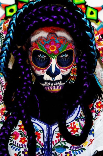 close up of a woman's head and shoulders wearing a Dia de Muertos costume at the festival in Oaxaca. Her face is painted like a skull with a brightly coloured pattern around the eyes and forehead and a pink flower in the middle of her forehead, and she is wearing thick black and purple plaits around her face as well as a white top embroidered with many small colourful flowers