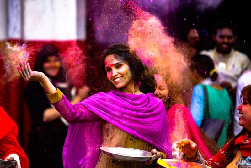 Indian woman in a purple shawl throwing colourful powder into the air in front of a crowd of people at Holi in India