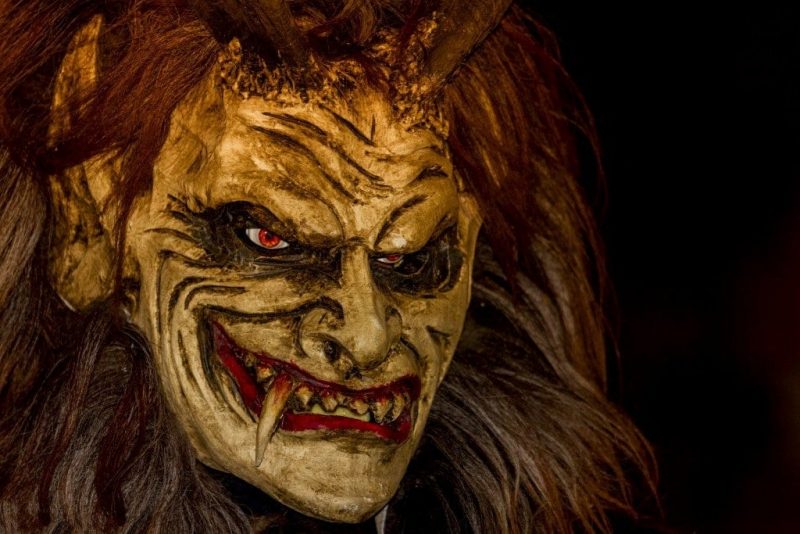 close up of a Krampus mask, a white demonic face with red lips, fangs, and narrowed eyes, surrounded by fur and long hair