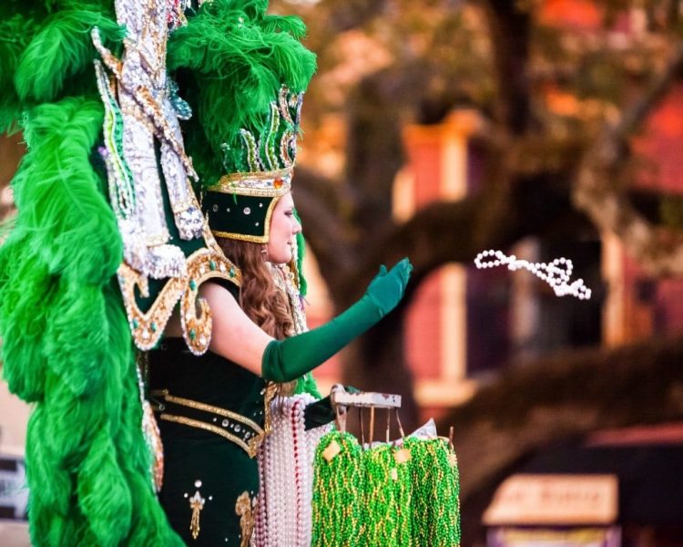 Woman in a black Mardi Gras costume with a large headress covered in diamante studs and green feathers throwing a white beaded necklace 