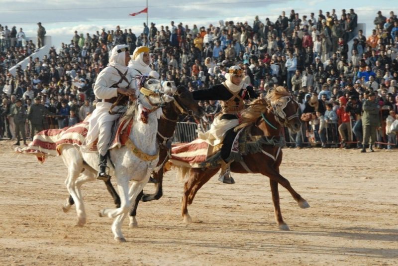 three men wearing traditional Saharan outfits with white shirts and trousers and white headscarves riding horses - one white horse and two brown - in a race on the desert sand with a large tiered stand of spectators behind