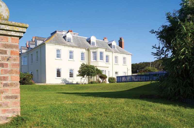 large white manor house with a grey tiled roof in a grassy lawn on a sunny day with clear blue sky behind