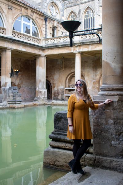Emily wearing a yellow long sleaved wrap dress and black leggings with black suede ankle boots and purple sunglasses standing with one hand on a large stone pillar in front of the green water of the ancient roman thermal pool in Bath, with the beige stone walls and pillars of the bathhouse behind. 