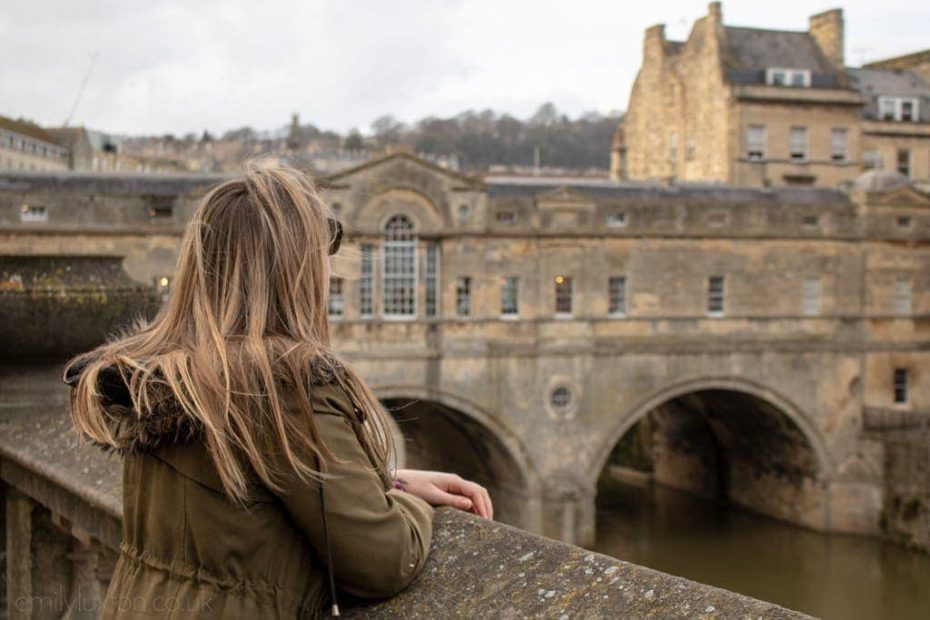 Emily wearing a khaki green parka with a fur lined hood with her long blonde hair down leaning against a low stone wall and looking out at Pulteney Bridge in Bath, a covered stone bridge lined with the backs of a terraced row of shops. Weekend in Bath.