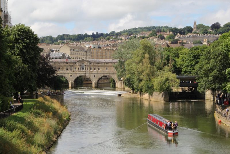 Wide river with a red canal boat sailing up it towards an arched stone bridge with the bath city skyline visible behind.