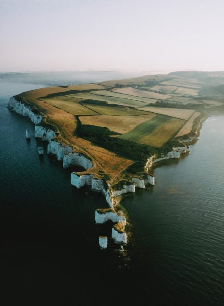 Aerial view of a peninsula with lots of fields over it and white chalk cliffs all the way around with several white chalk rock stacks leading away from the tip into the sea - on the Isle of Purbeck Dorset England