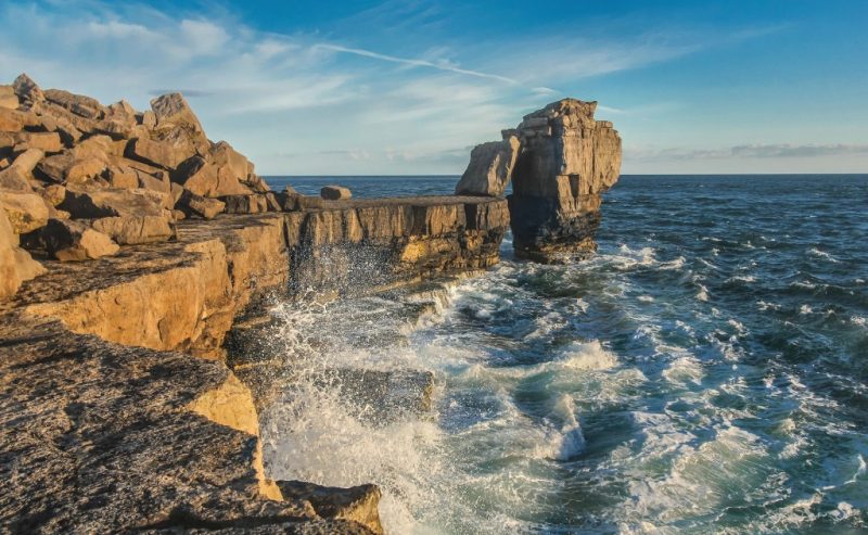 Pulpit Rock on Portland, a large grey rock stack in the sea besides the low rocky cliff edge with small waves crashing against the rocks in the foreground