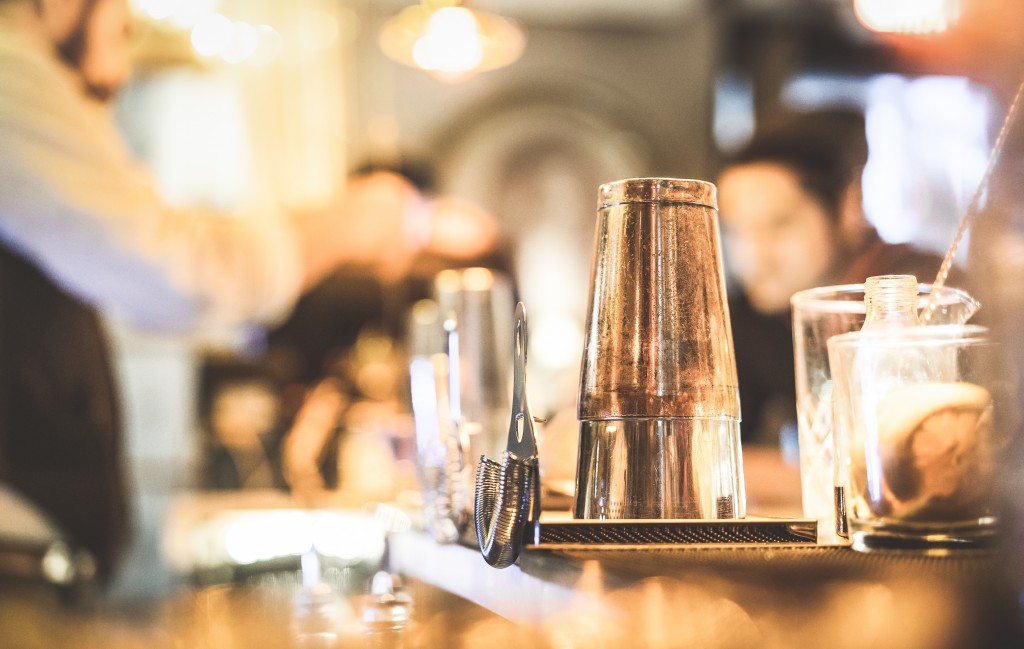 close up of a metal cocktail shaker and muddler next to some glasses on a bar with the background out of focus 