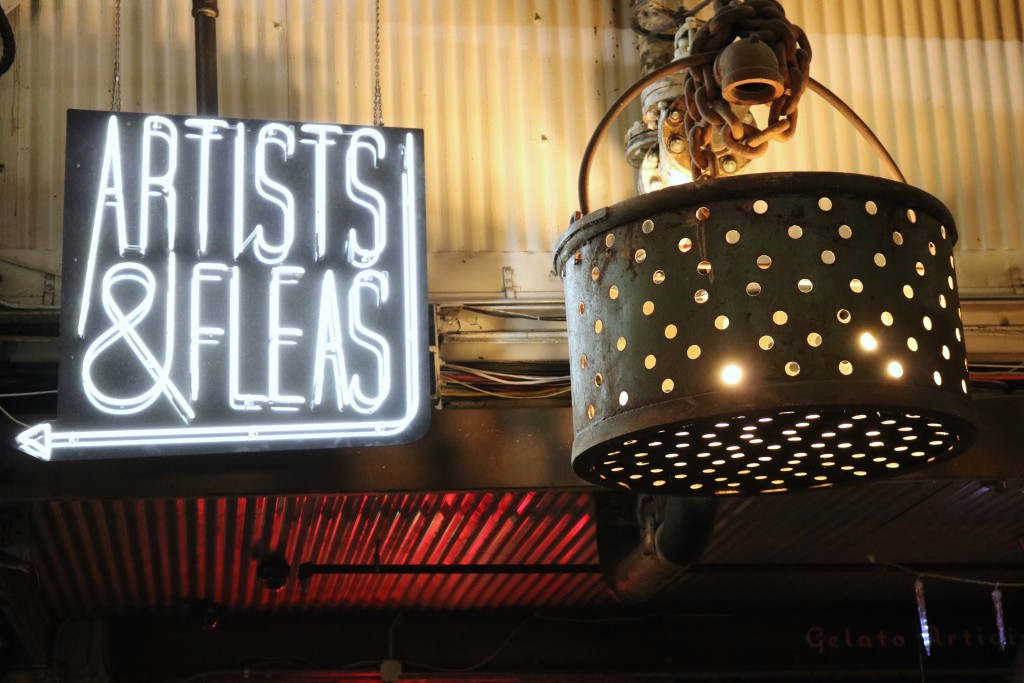 neon sign which says artists & fleas in white writing on a black background next to a metal light made from a bucket with holes in hanging from a corrugated iron cieling - cool things to do in nyc