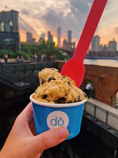 close up of a hand holding a blue paper tub with cookie dough icing in front of the new york city skyline at sunset - foodie highlights NYC