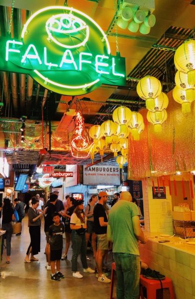 crowded indoor market hall with food stalls in a row - the closest one is decorated with paper lanterns and has a large green neon sign for falafel - New York food hotspots