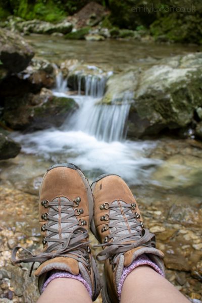 A pair of feet wearing brown leather hiking shoes and pink wool socks in front of a small waterfall over a rock in a river