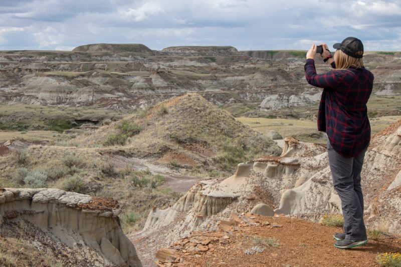 Emily standing on a small orange cliff overlooking a valley filled with wild grass and brown rock formations with low grey-brown cliffs on the far side topped with wild grass. She is wearing grey shoes, grey trousers, and a blue and red tartan-style long sleeved shirt and a black baseball cap and holding her camera up to take a photo. Solo road trip in Alberta.