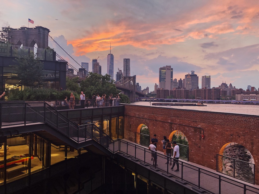 new york city at sunset with people on the roof terrace of an apartment building in the foreground and the city skyline and brooklyn bridge behind