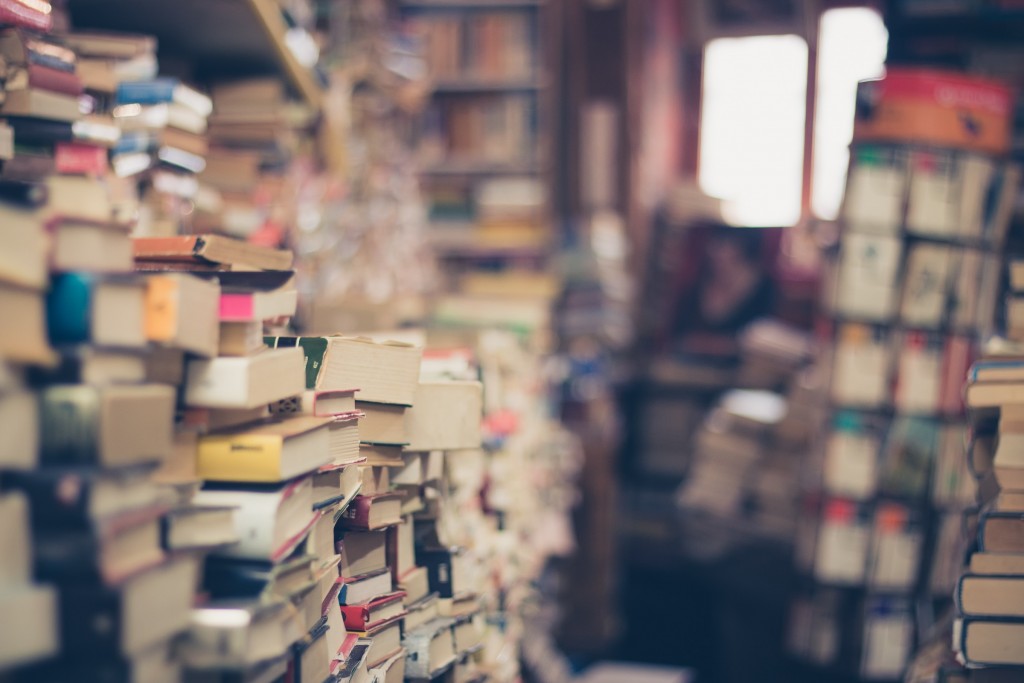 cluttered book store filled with many stacks of books with a window out of focus in the background