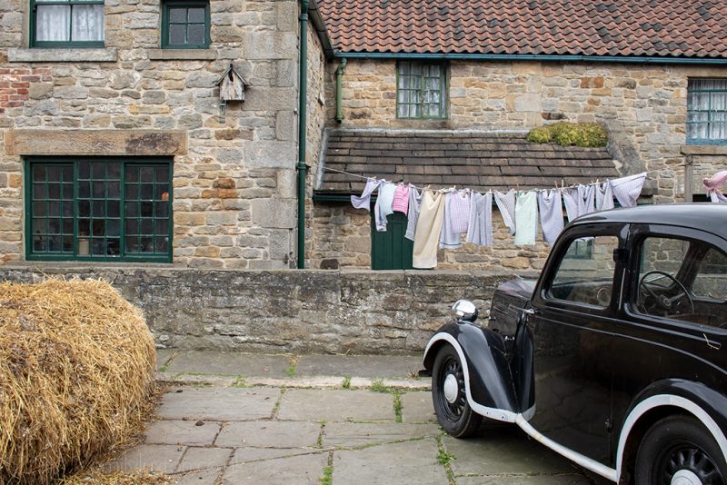 Historic stone cottages with red tiled roods and green wood frames around the windows, there is a haybale in front and a 1950s style black car parked on the flagstones outside at Beamish Museum in County Durham