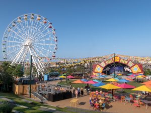 Colorful amusement park at Dreamland Margate with a Ferris wheel, stage, tables under umbrellas, and a pizza stand. People are walking and sitting, enjoying the lively atmosphere.
