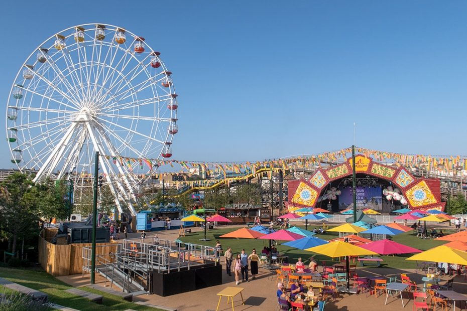 Colorful amusement park at Dreamland Margate with a Ferris wheel, stage, tables under umbrellas, and a pizza stand. People are walking and sitting, enjoying the lively atmosphere.