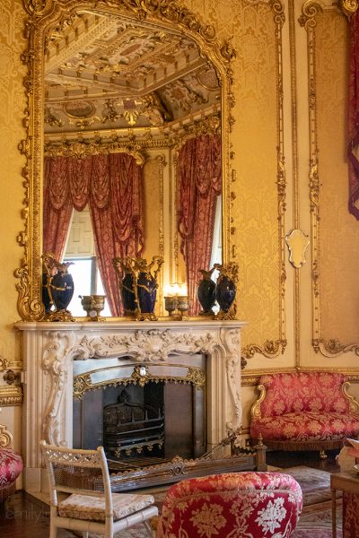 Interior of Raby Castle with an elaborately furnished room with a carved white fireplace, large mirror with gold frame, and golf wallpaper. there are small red and gold armchairs around the fireplace. 