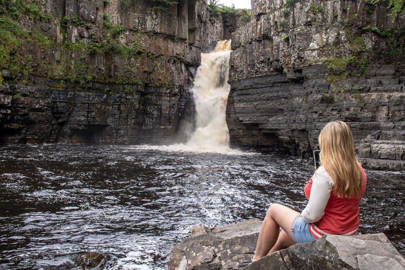 Emily sitting with her back to the camera wearing a cream and red jumper and denim shorts facing a large waterfall running down a stark grey cliff - High Force Falls Durham Dales