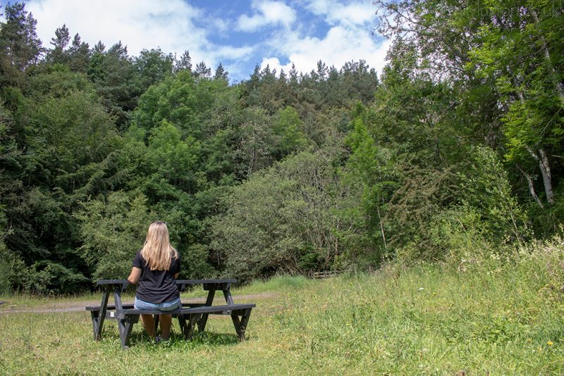 Emily wearing denim shorts and black t shirt sat at a black picnic bench in a grassy clearing surrounded by trees. Things to do in county durham uk