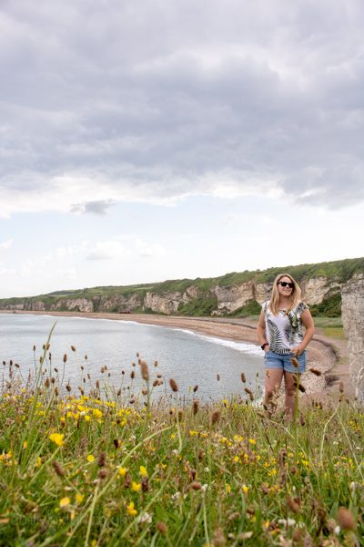 Emily wearing a white top with palm leaves printed on it and denim shorts walking with her hands in her pockets across a wildflower meadow at the top of a low cliff with a view of a small bay and a shingle beach behind her and the Durham Heritage Coast beyond that. 