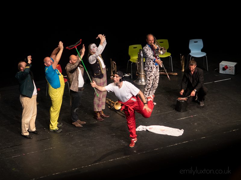 Group of people dressed as clows or wearing heavily patterned suits dancing on a stage. One man is playing trumpet and the man in the front is wearing bright red trousers and a white t shirt and holding a plastic green broom in the air. Things to do in durham city