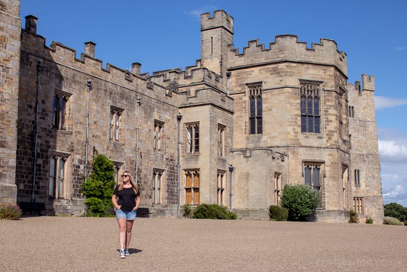 Emily wearing denim shorts and black t shirt walking with her hands in pockets away from a large stone castle with turreted roof. Things to do in County Durham UK