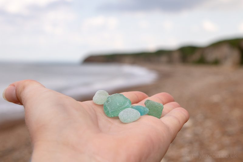 Close up of a hand holding lots of pieces of green and blue seaglass with the beach and cliffs out of focus behind. County Durham Heritage Coast