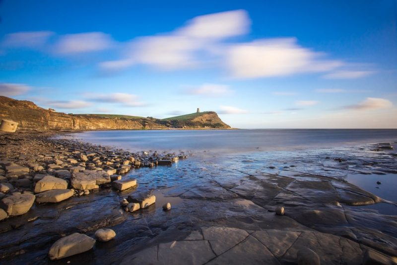 wide bay with flat grey rocks in the foreground and a low grassy headland across the bay with a small stone tower on top. UK staycation ideas. 