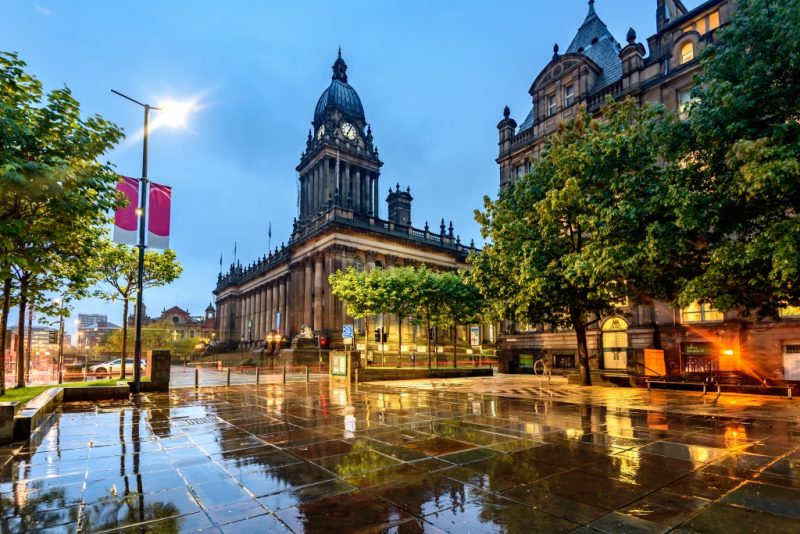 Exterior of Leeds Town Hall at night, a historic stone building with a domed clock tower in a square with leafy green trees around, all reflected in puddles on the flagstone road 