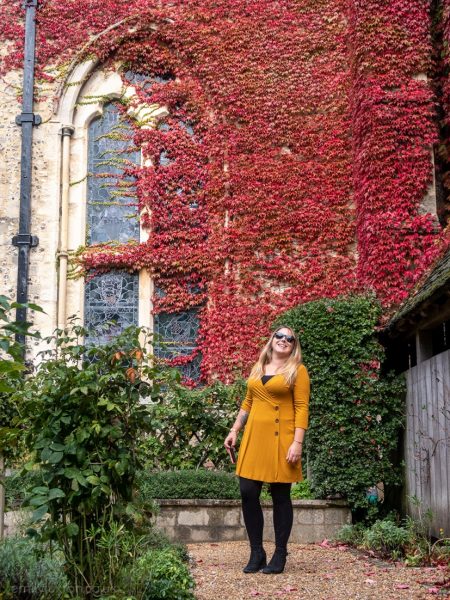 Emily wearing a mustard yellow wrap dress, black leggings, and black ankle boots, standing in a garden in front of the stone wall of Winchester Great Hall which is covered in red ivy because it is Autumn.