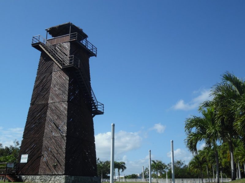 wooden airport tower with a staircase around the outside of the square tower against a blue sky with palm trees in front at Cancun Airport in Mexico