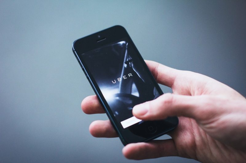 Hand holding a smartphone with the Uber homescreen on it and a grey background behind the phone. 