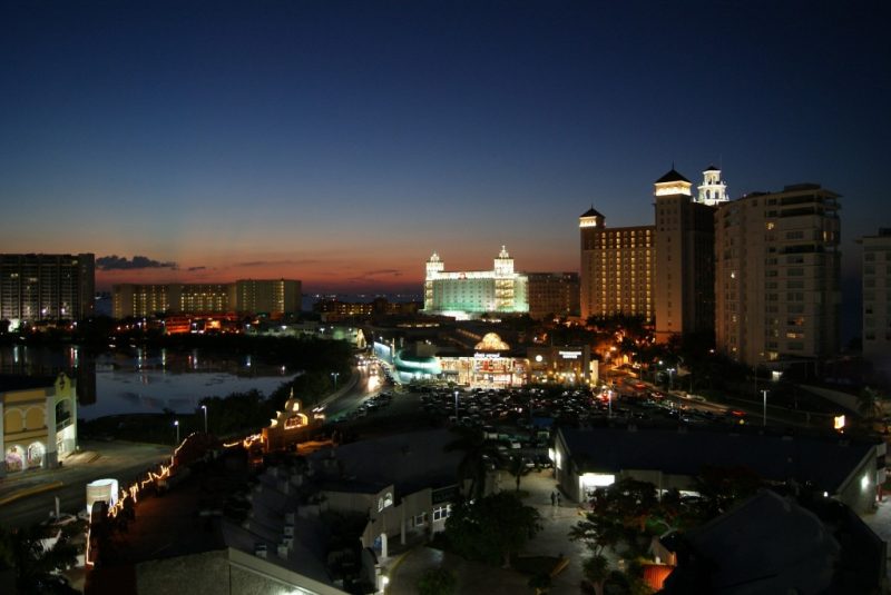 shot of Cancun Hotel strip at night with several highrise hotels and brightly lit buildings along a road next to a lagoon
