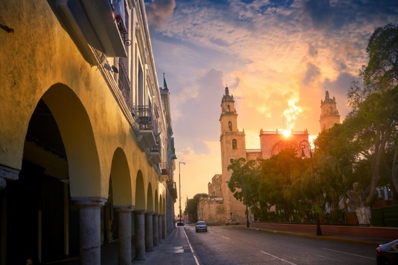 a yellow building with arches along the edge of the street with a park on the right side of the road and a church with a sunset sky behind that - what to do in Merida Mexico