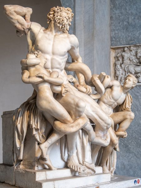 white marble statue in a museum of a nude man with curcly hair and beard. there are two smaller nude figures on either side of the man and a snake wrapped around them all. 