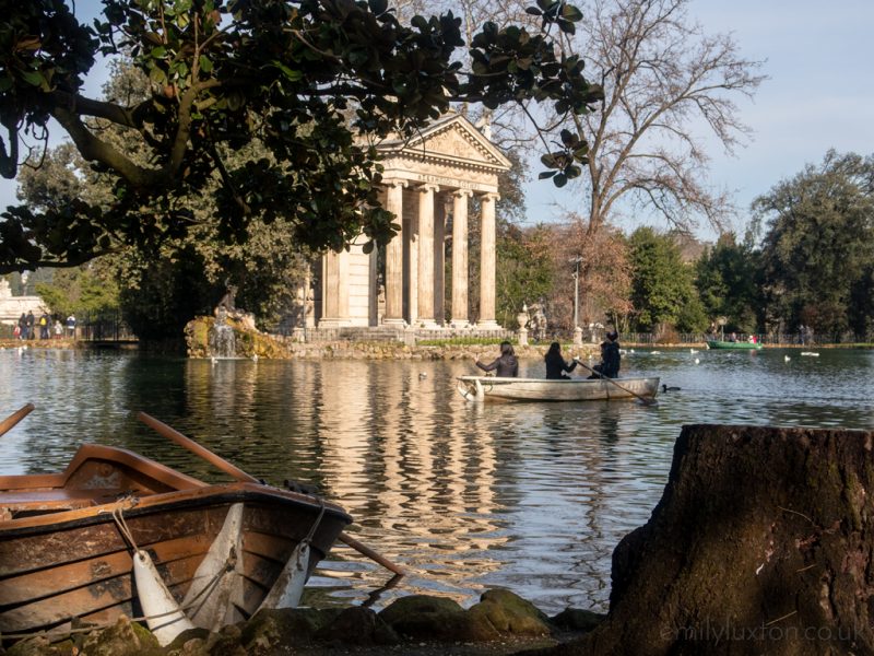 A small lake in the Borghese Gardens surrounded by trees with three people in a wooden rowing boat going past a small island with a white stone temple with four pillars holding up a triangular roof