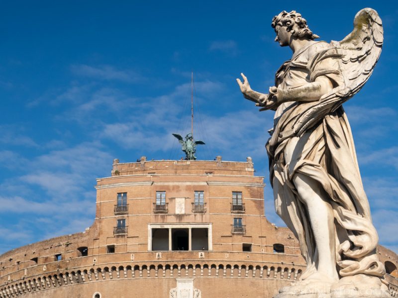 large white marble statue of an angel with wings and long flwoing robes holding up one hand in front of a large circular building of orange coloured bricks