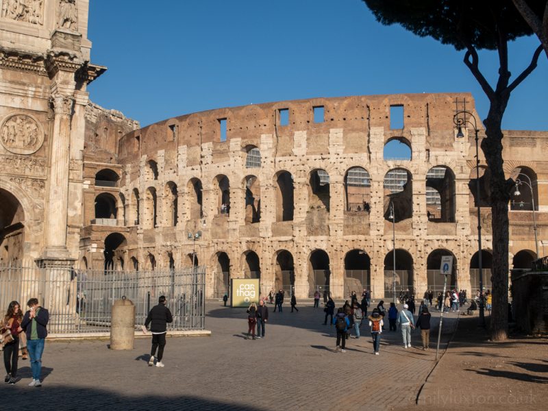 exterior of the Colosseum in Rome with a few people walking in the square in front and clear blue sky overhead
