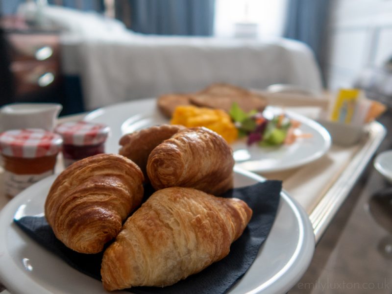 Room service breakfast with four croissants on a white china plate on a tray with more food and drinks out of focus behind and the hotel bed vicible behind that. 