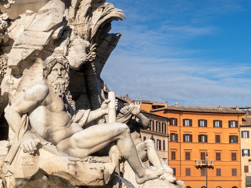 White stone statue of a nude man with a long beard in front of several tall orange apartment buildings on a sunny day - Rome in January