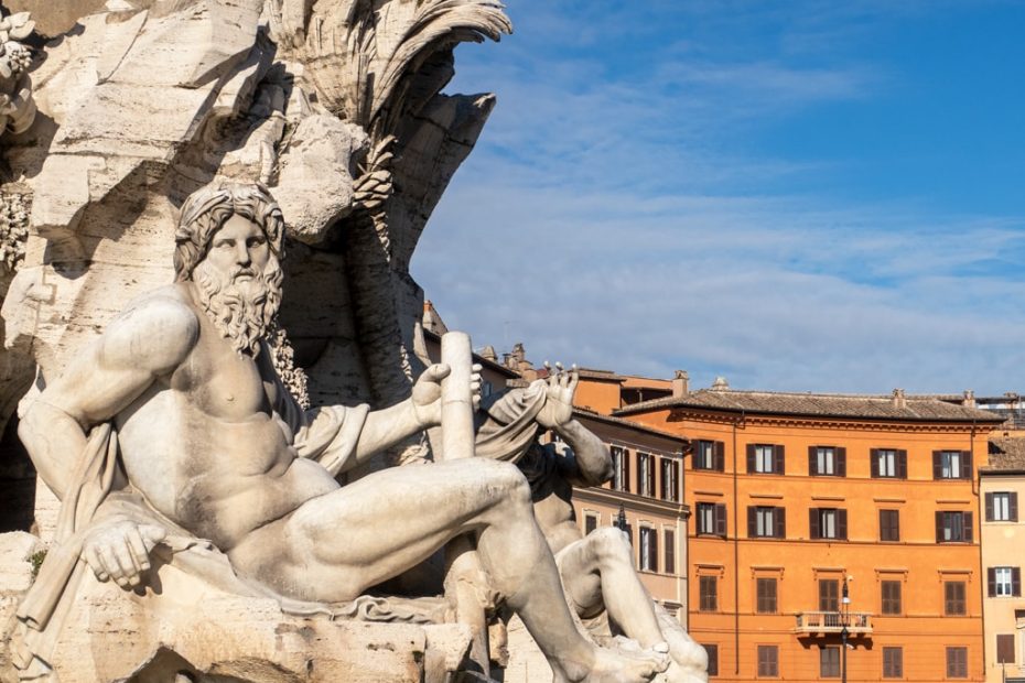 White stone statue of a nude man with a long beard in front of several tall orange apartment buildings on a sunny day - Rome in January